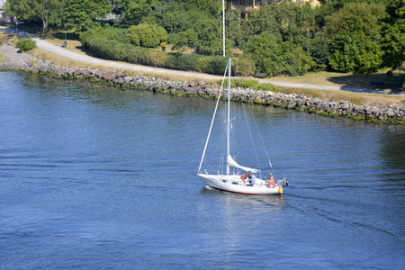 Baltic sea, Sweden seashore. July, 25, 2014. A white sailboat sailing along the strait in clear weather. Green plants cover the shore.のeditorial素材