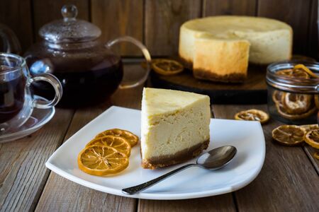 A slice of cheesecake on a white plate with lemon slices and a spoon. In the background there is a glass teapot with hot tea and a Cup of teaの写真素材
