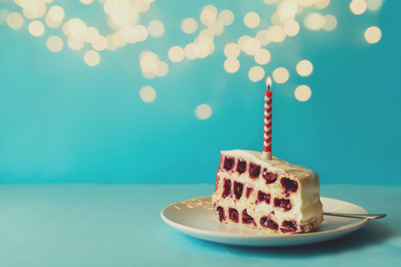 Slice of white birthday cherry cake with colorful candles on blue background against defocused light. Holiday celebration concept. Copy spaceの写真素材