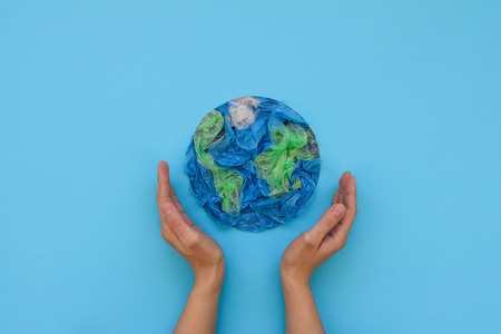 Hands holding planet Earth made from plastic disposable packages on blue background. Save the world,...