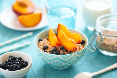 Porridge bowl oatmeal with peach and blueberries on turquoise background. Soft focus. Healthy eating concept.の写真素材