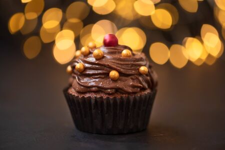 Chocolate cupcake with golden candies on dark wooden background against blurred lights. Selective focus. Unhealthy food.の写真素材