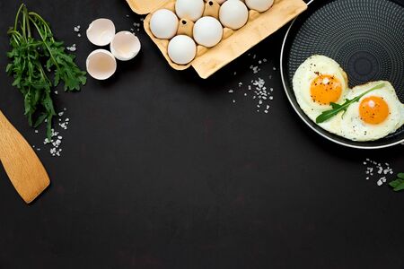 Fried eggs in frying pan with arugula leaves, salt and pepper for breakfast on black wooden...