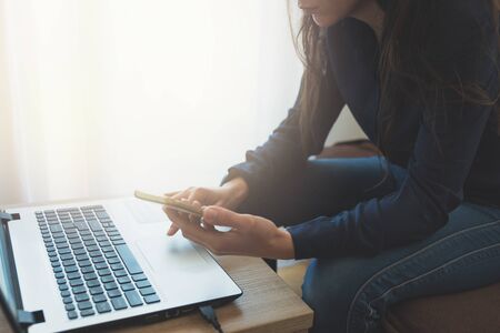 Young adult  touching smartphone sitting near laptop. Soft focus. Job at home. Female freelancer.の写真素材