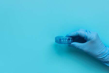 Hand of medic wearing blue latex glove holding myofunctional trainer on blue background. Teeth...