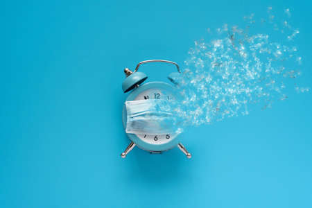 Alarm clock wearing into protective mask with dispersion effect on blue background. Quarantine or covid-19 concept. Top view. Flat layの写真素材