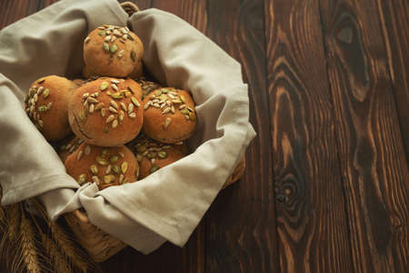 Basket of fresh homemade crispy bread buns with bran, sunflower and pumpkin seeds on brown wooden background. Top view.の写真素材
