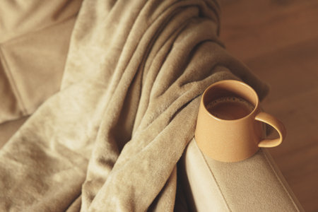 Mug of hot coffee stay on arm of chair by blanket