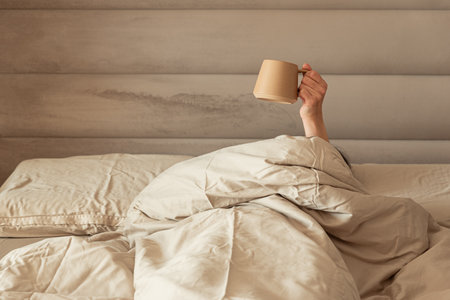 A hand holding a coffee mug emerging from a cozy bed.の写真素材