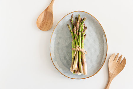 Fresh asparagus tied bundle on plate and wooden utensils on white table. Top viewの写真素材