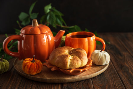 Autumnal tea set with pumpkin-shaped teapot and mug, and a baked bun on a wooden table.の写真素材