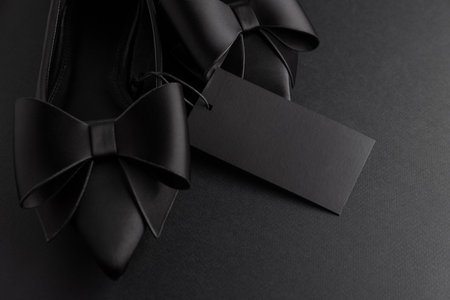 Elegant black pointed-toe shoes adorned with bows and a blank tag on black backgroundの写真素材