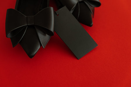 Elegant black pointed-toe shoes adorned with bows and a blank tag on red backgroundの写真素材