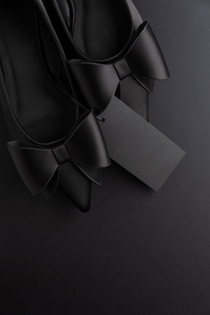 Elegant black pointed-toe shoes adorned with bows and a blank tag on black backgroundの写真素材