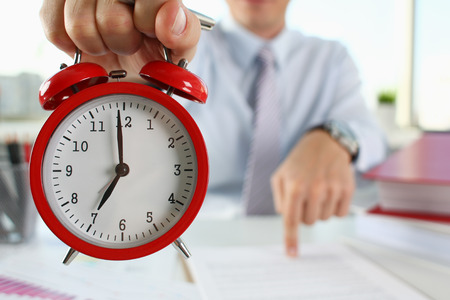 Male hand on the alarm clock a red color stands in the office on the table showing seven oclock in...