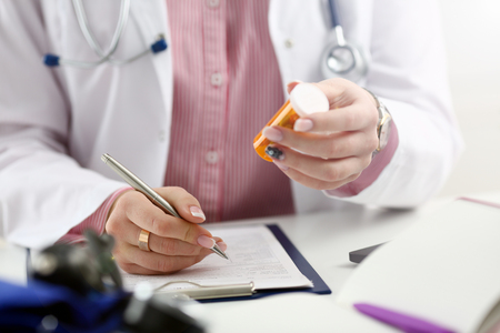 Female medicine doctor hand hold jar of pills and write prescription to patient at worktable. Panacea and life save, prescribing treatment, legal drug store concept. Empty form ready to be usedの写真素材