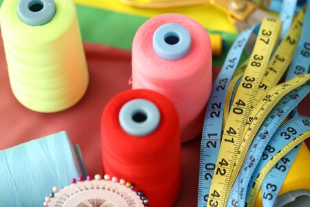 Focus on multicolored spools of thread lie on table near measuring tapes and special needles covered in textile and fabric of various colors and qualities. Fashioner market concept. Blurred backgroundの写真素材