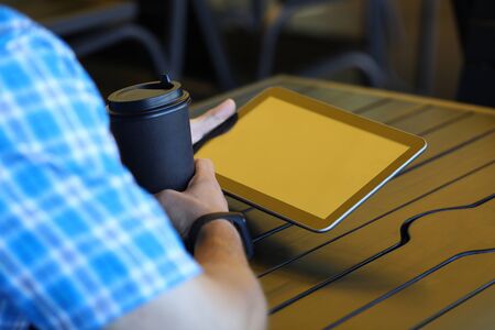 Focus on male hands holding modern tablet with blank display. Man sitting at wooden table in cafe. Guy drinking coffee and using high-tech gadget. Copy space on screen. Blurred backgroundの写真素材
