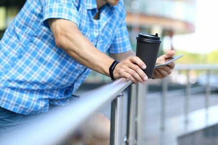 Focus on male standing near handrail and reading news on tablet. Smart guy in fashionable shirt holding coffee cup and enjoying lunchtime. Blurred backgroundの写真素材