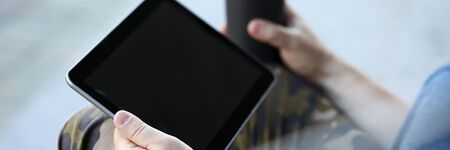 Focus on male hands holding stylish gadget and paper cup of coffee in hands. Guy surfing internet,...