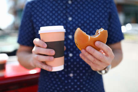 Man holds glass with hot coffee and hamburger in his hand