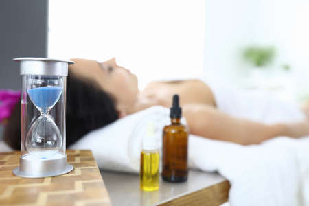 Woman lies in spa salon next to an hourglass and aromatic oils