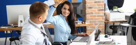 Disabled businessman sits in wheelchair at work table in office and greets businesswoman.の写真素材
