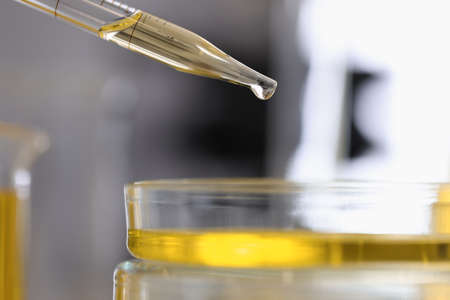 Dispenser filled with yellow chemical liquid pours drop into a glass dish closeupの写真素材