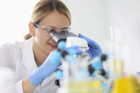 Woman in rubber gloves and protective chemical glasses looks through microscope in laboratory portraitの写真素材