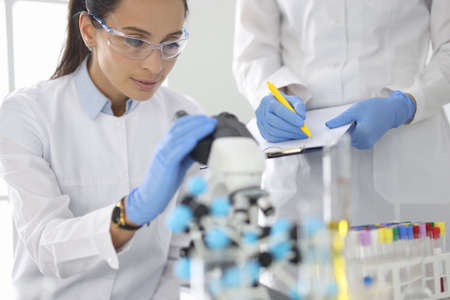 Young woman in white uniform wearing protective chemical goggles and rubber gloves looks into microscopeの写真素材