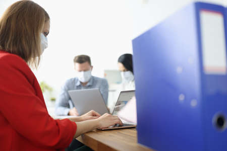 Young woman in business suit and masked face is typing on laptop keyboard in office with colleagues on backgroundの写真素材