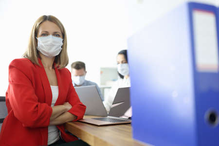 Business woman in red suit and protective mask on face sits at table in officeの写真素材