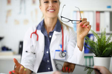 Woman doctor is sitting at table smiling and holding glasses in her handsの写真素材