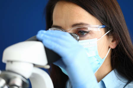 Woman in protective medical mask with glasses and gloves looks through a microscopeの写真素材