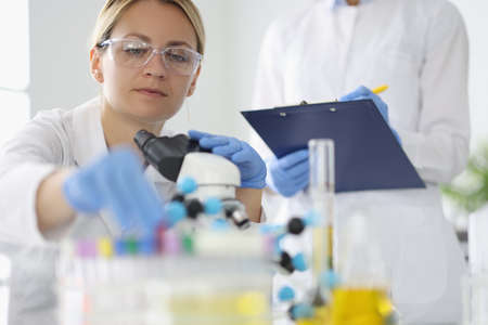 Researcher with glasses takes test tube on table with microscope near colleague writes down the results.の写真素材