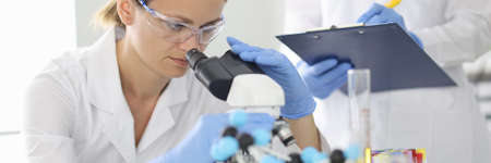 Woman scientist in protective glasses looks through microscope and trains assistant in chemical laboratoryの写真素材