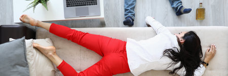 Woman lies on couch next to an alcohol bottle and man lies.の写真素材