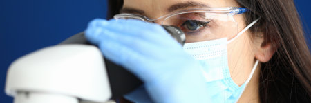 Woman in protective medical mask with glasses and gloves looks through a microscopeの写真素材