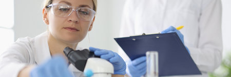 Researcher with glasses takes test tube on table with microscope near colleague writes down the results.の写真素材