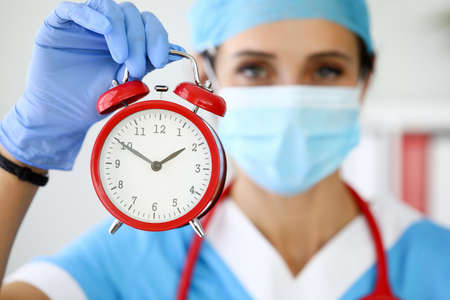 Woman doctor in protective medical mask holding red alarm clock in her hands