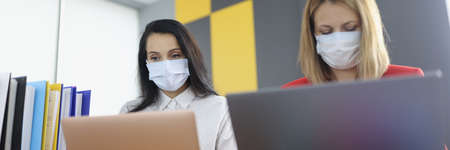 Two women at desk in protective medical masks work on laptops.の写真素材