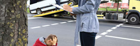 An agent fills out documents after car accident, driver sits nearby and holds his head with his handsの写真素材