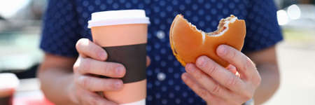 Man holds glass with hot coffee and hamburger in his handの写真素材