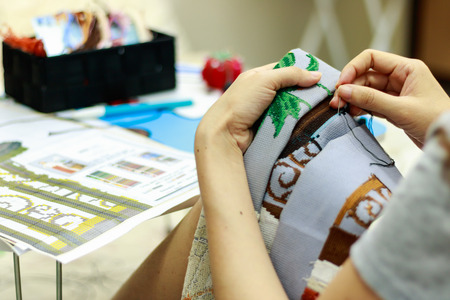 Young woman doing cross stitch at home. Close up of embroidery. Cross stitch is a hobby.