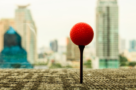 Orange golf ball on tee behind cityscape scene in the office. Go out of ofice.の写真素材