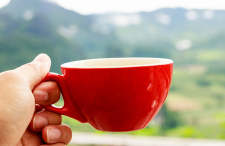 Hand holding a red cup of coffee with blurred mountain background in morning time.の写真素材