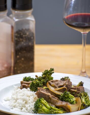 Cooked beef and broccoli stirfry served on rice with a wine glass and spices in the backgroundの写真素材