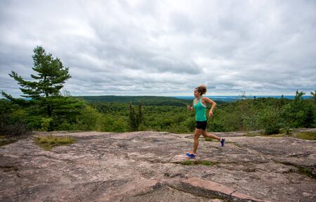 A young woman wearing fitness clothing running across rocks with forest background