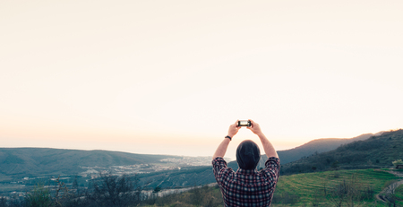 A young man standing atop a mountain at sunset takes pictures of the skyの写真素材