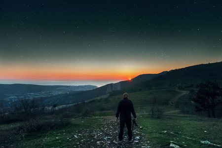 A young man stands on top of a mountain and enjoys the sunset and starry skyの写真素材